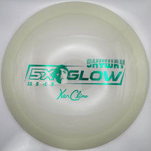 A translucent pale yellow disc golf disc with green printed graphics, including 'SHIMURA GLOW' and 'Xa Clime', resting on a white foam insert.