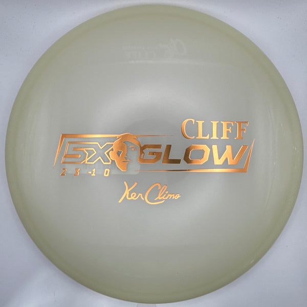 A disc golf disc, specifically a Cliff Glow X2 from Yakucho, displayed inside its white presentation box. The disc is a pale, translucent cream color with metallic gold foil stamping.