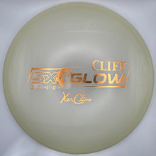 A disc golf disc, specifically a Cliff Glow X2 from Yakucho, displayed inside its white presentation box. The disc is a pale, translucent cream color with metallic gold foil stamping.