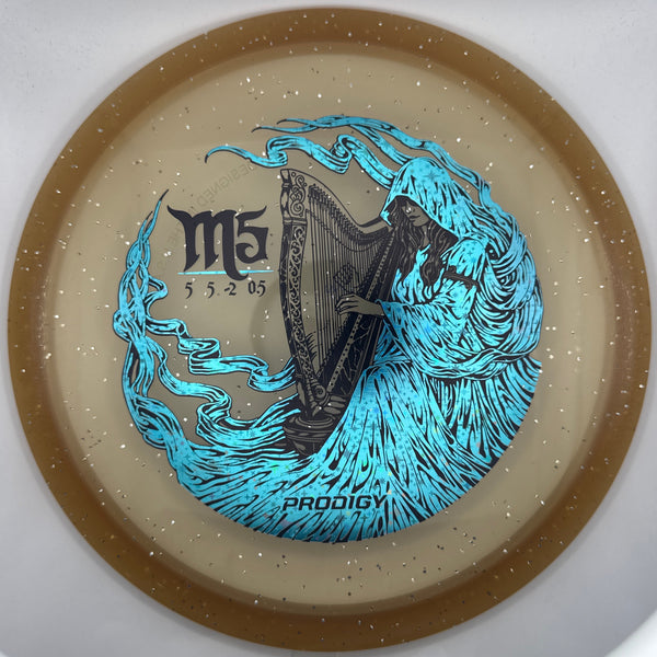 A Prodigy M5 disc golf disc with a speckled beige plastic body, featuring a brown outer rim and a detailed graphic of a harp surrounded by blue flames. The Prodigy brand name is printed at the bottom of the graphic.