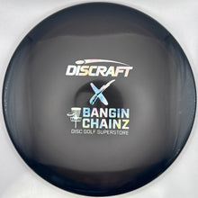 A close-up of a Discraft x Bangin' Chainz disc golf disc, featuring a dark gray/black body with white and light blue branding, mounted on a white cardboard backing with a purple edge visible.