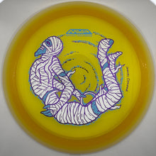 A yellow Discraft flying disc featuring a stylized white mummy illustration with blue and purple details, photographed inside a circular opening.