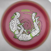 A pink disc golf disc with a green and white mummy graphic, photographed inside a circular white presentation box.