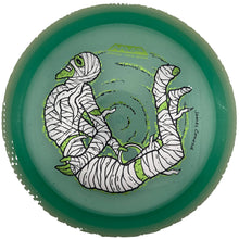 A translucent green disc golf disc featuring a cartoon mummy design with white bandages and lime green accents, photographed from directly above inside a circular opening.