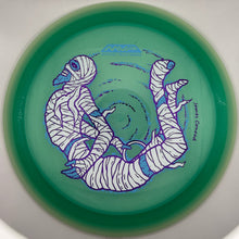 A green disc golf disc featuring a stylized mummy design in white and blue, photographed from directly above inside a white circular frame.